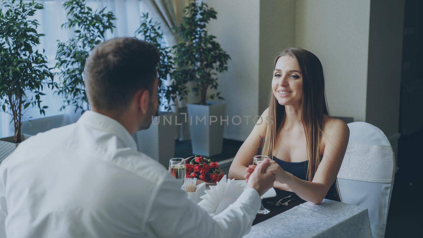 Young attractive girl in fancy clothes is talking to her bearded boyfriend while dining in restaurant. Flowers, champagne glasses, green plants and tableware are visible.