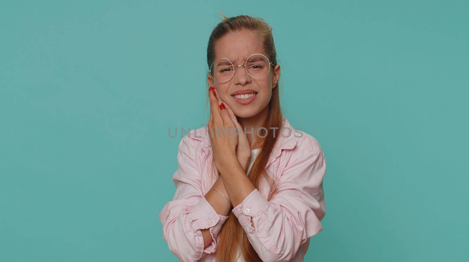 Dental problems. Pretty teen young girl touching cheek, closing eyes with expression of terrible suffer from painful toothache, sensitive teeth, cavities. Student child kid on blue background indoors