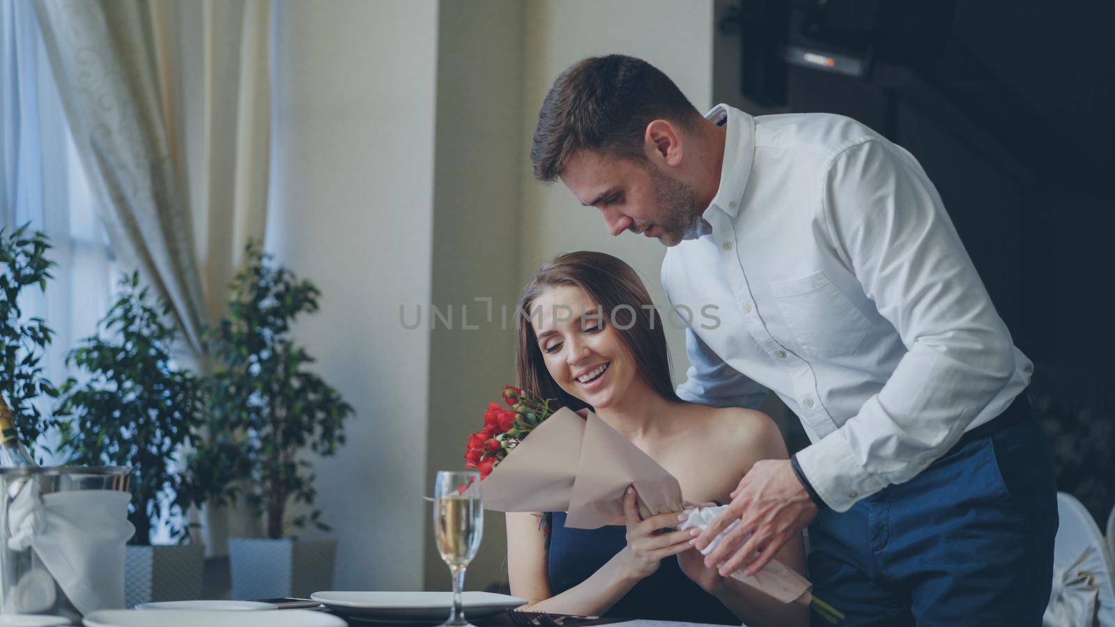 Beautiful woman is waiting for her boyfriend in restaurant, he is coming and giving her flowers, kissing her. Happy girl is smelling flowers and smiling with pleasure. by silverkblack