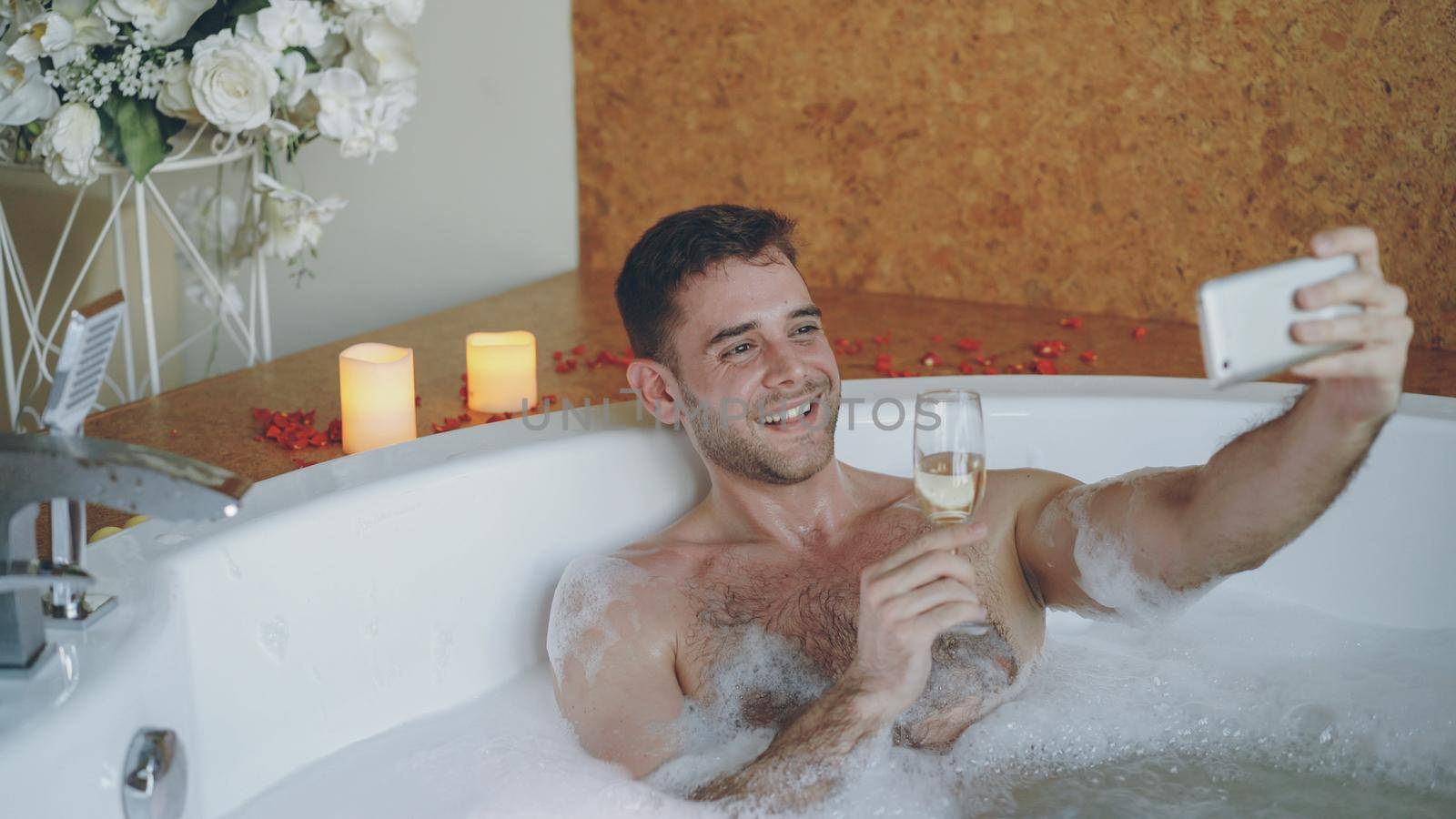 Handsome young man is taking selfie with champagne glass using smartphone in hot tub in modern spa salon. He is smiling and posing looking at camera. by silverkblack
