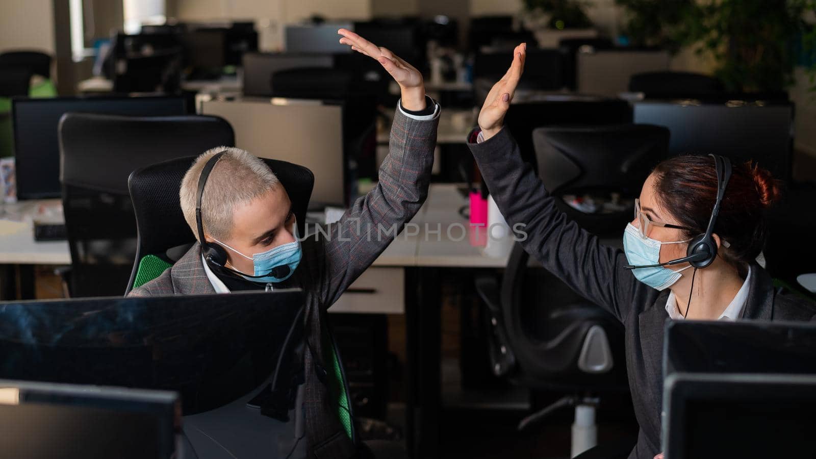 Two business women in masks are giving a high five while sitting at one desk in the office.