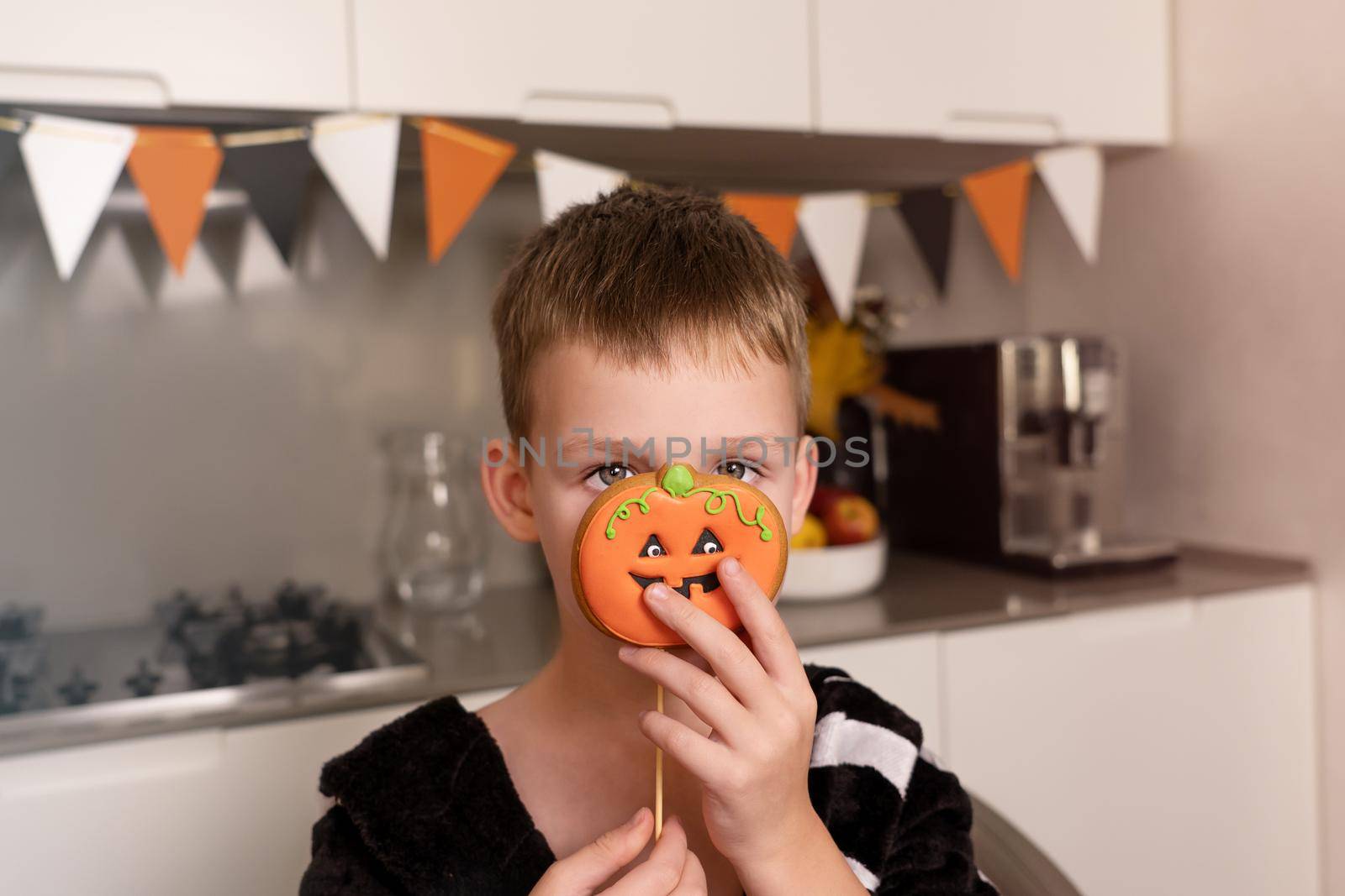 Little boy, Caucasian in Halloween style. the child is holding a pumpkin-like gingerbread cookie with a funny face near the eyes. Against the backdrop of home cooking. Home baking concept