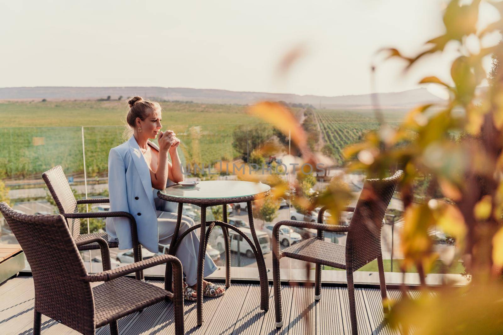 A middle-aged woman sits in a street cafe overlooking the mountains at sunset. She is dressed in a blue jacket and drinks coffee admiring the nature. Travel and vacation concept