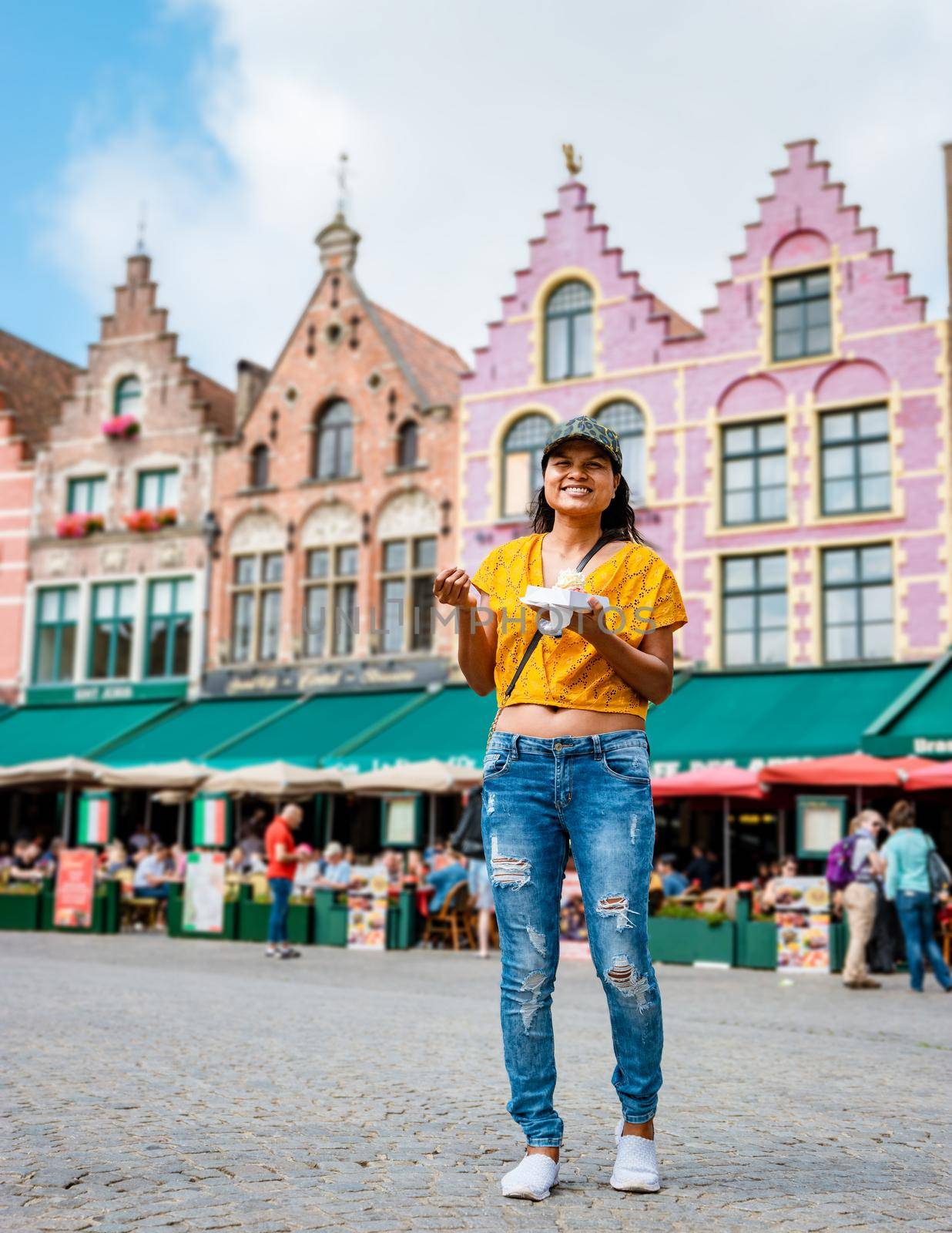 Women on a city trip in Brugge eating a Belgium waffle, the colorful house in the old city of Brugge with restaurants at the square.