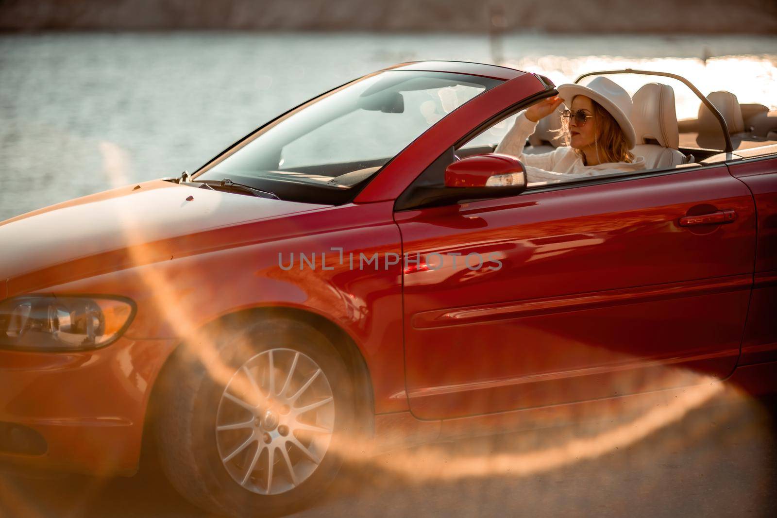 Outdoor summer portrait of stylish blonde woman driving red car convertible. Fashionable attractive woman with blond hair in a white hat in a red car. Sunny bright colors taken outdoors against the sea. by Matiunina