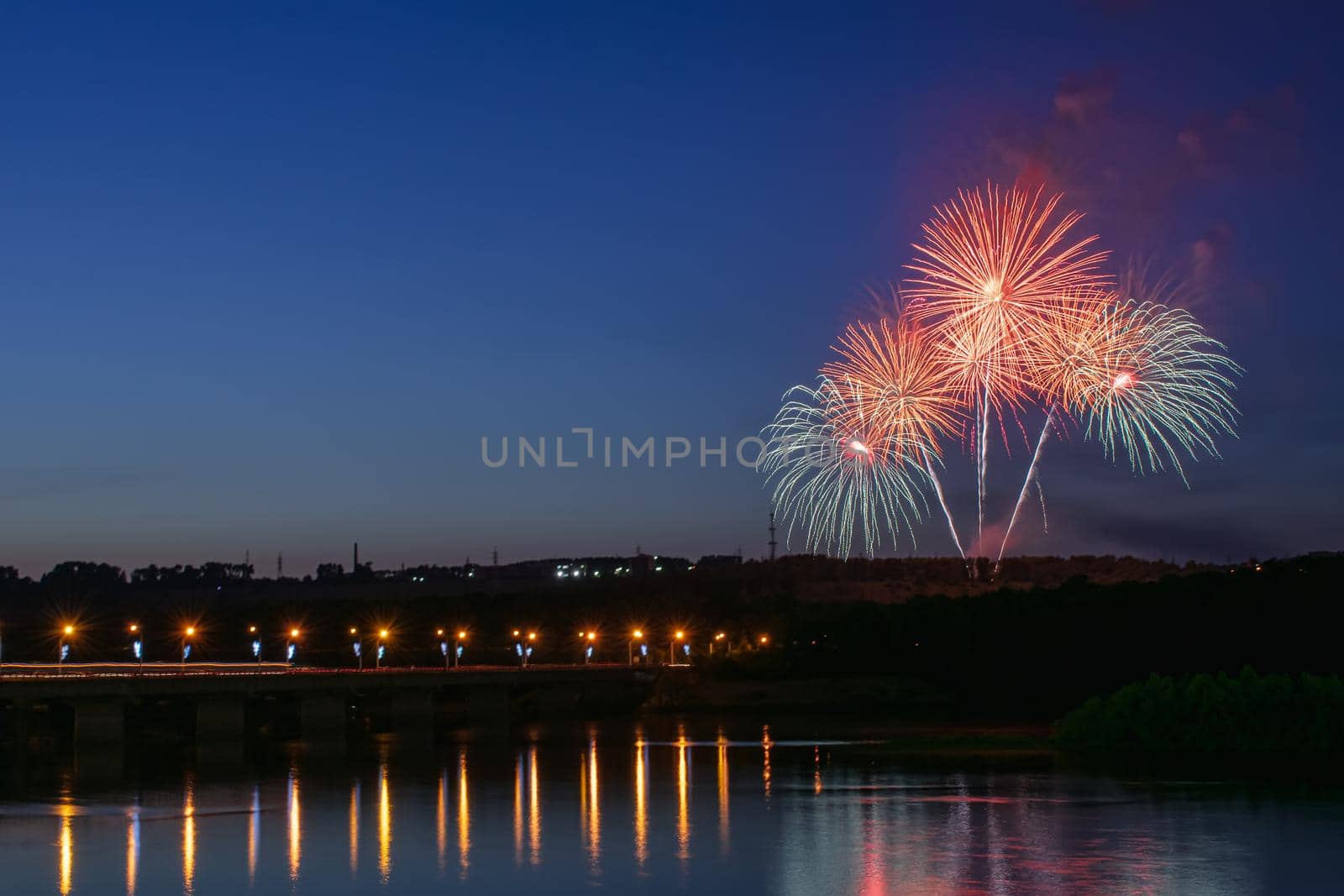 Fireworks against the background of a clear night sky near the river in which the light is reflected and the bridge across this river
