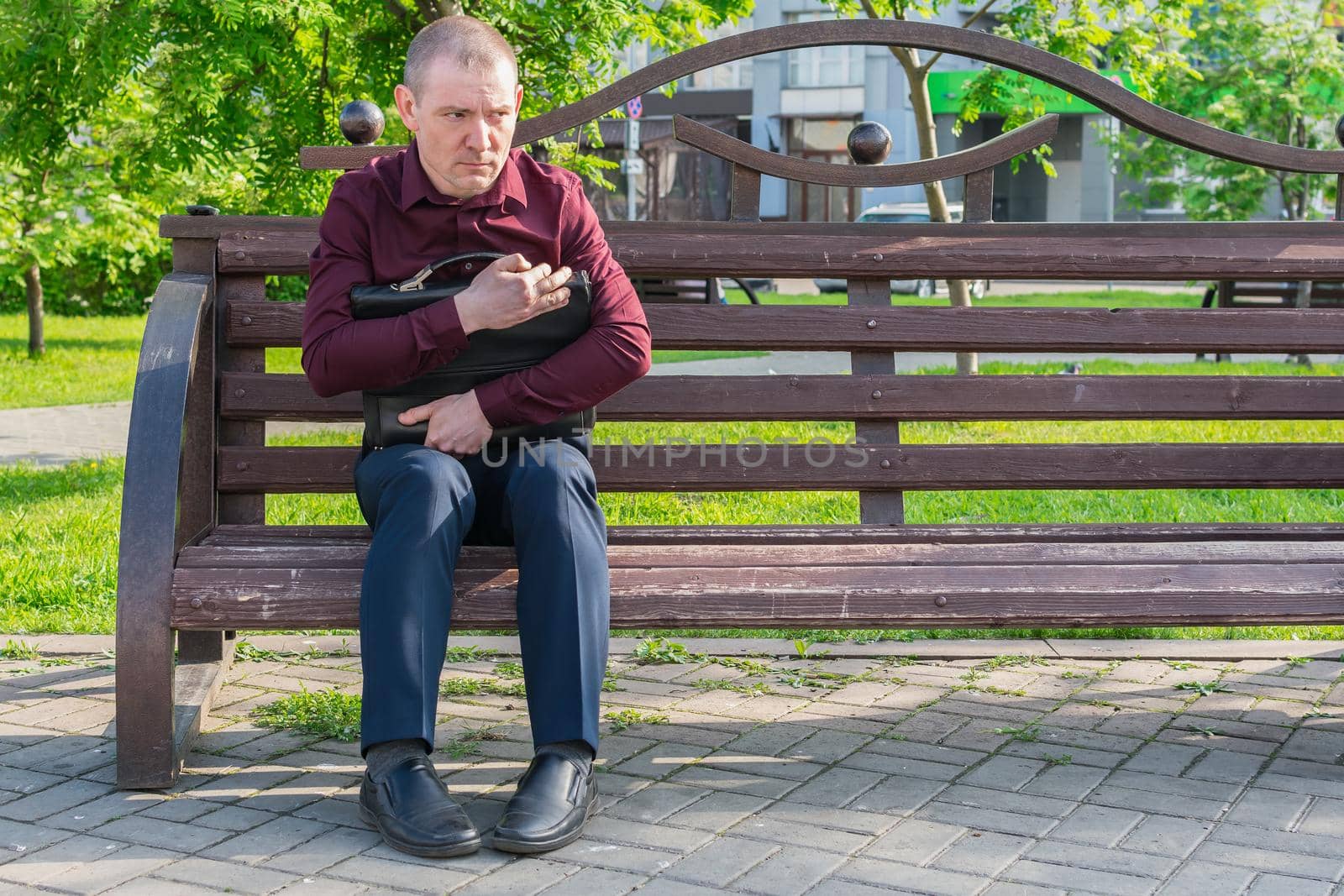 A scared man with a suitcase and a suspicious look sits on a park bench, looking like a spy or an office worker