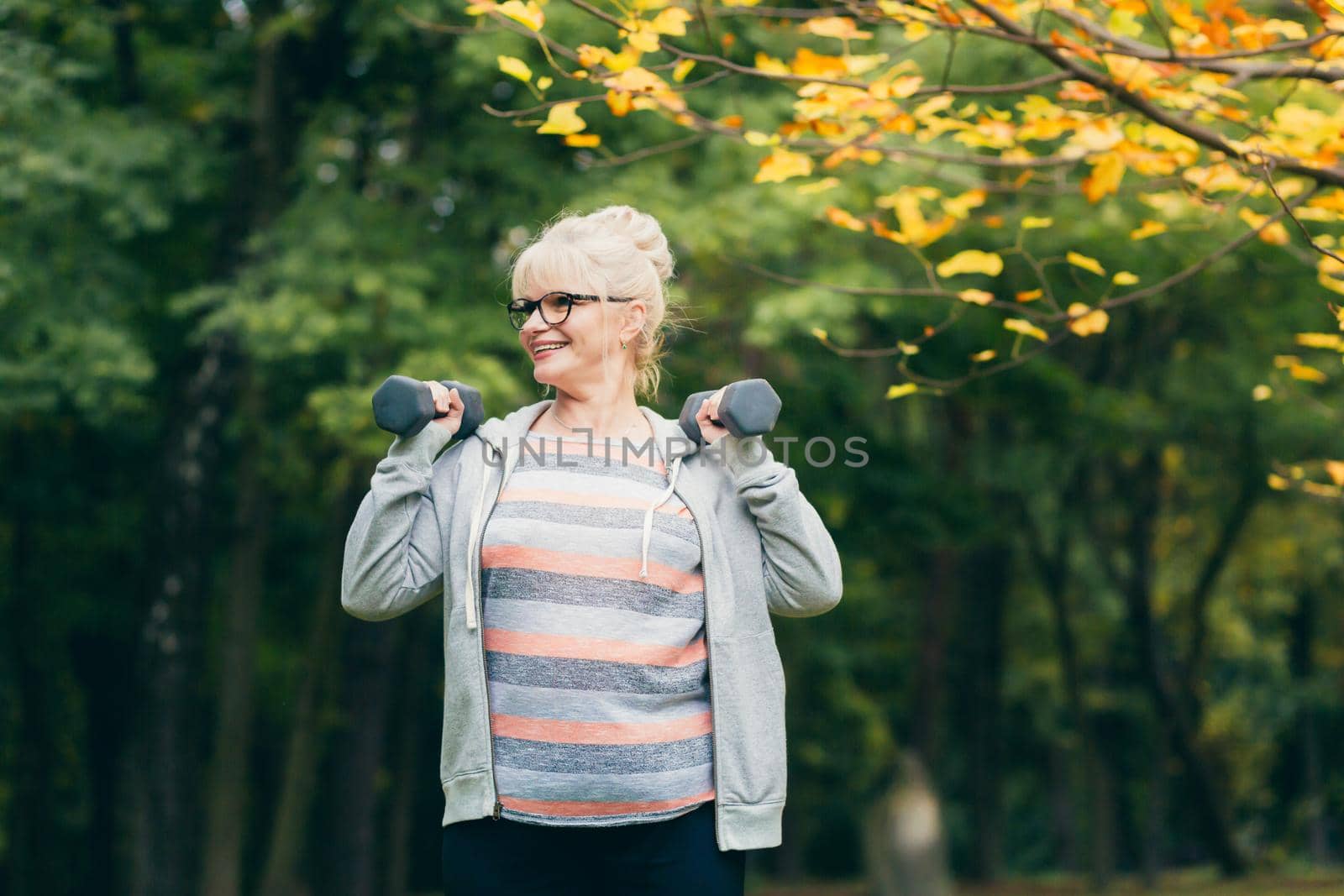 Senior woman standing and doing sports exercises with dumbbells in her hands in the park on the grass, exercising, leading a healthy active lifestyle sitting on the grass