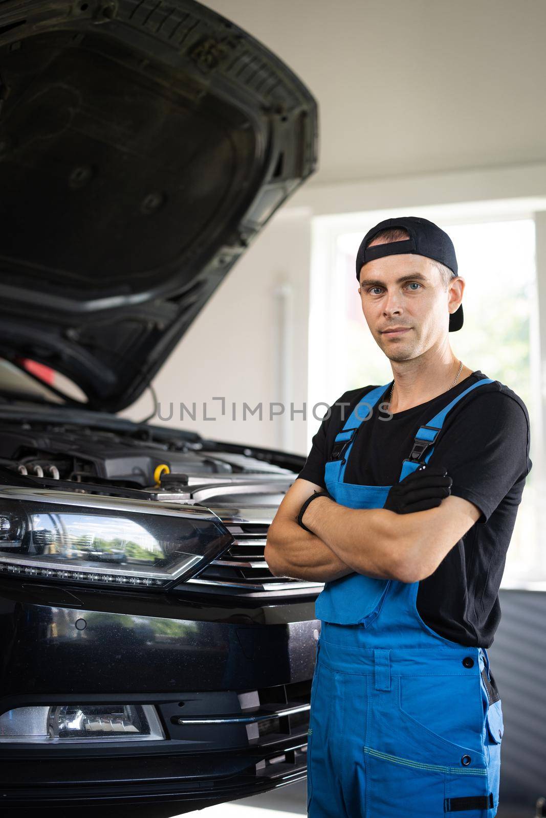 Portrait shot of caucasian man in blue uniform and hat standing in big auto garage, looking to camera and crossing hands. Pretty male mechanic in maintaining service by uflypro