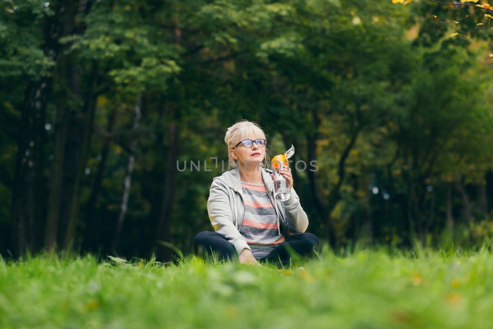 Senior beautiful woman sitting in the park on the grass and drinking fresh juice, water after sports, yoga