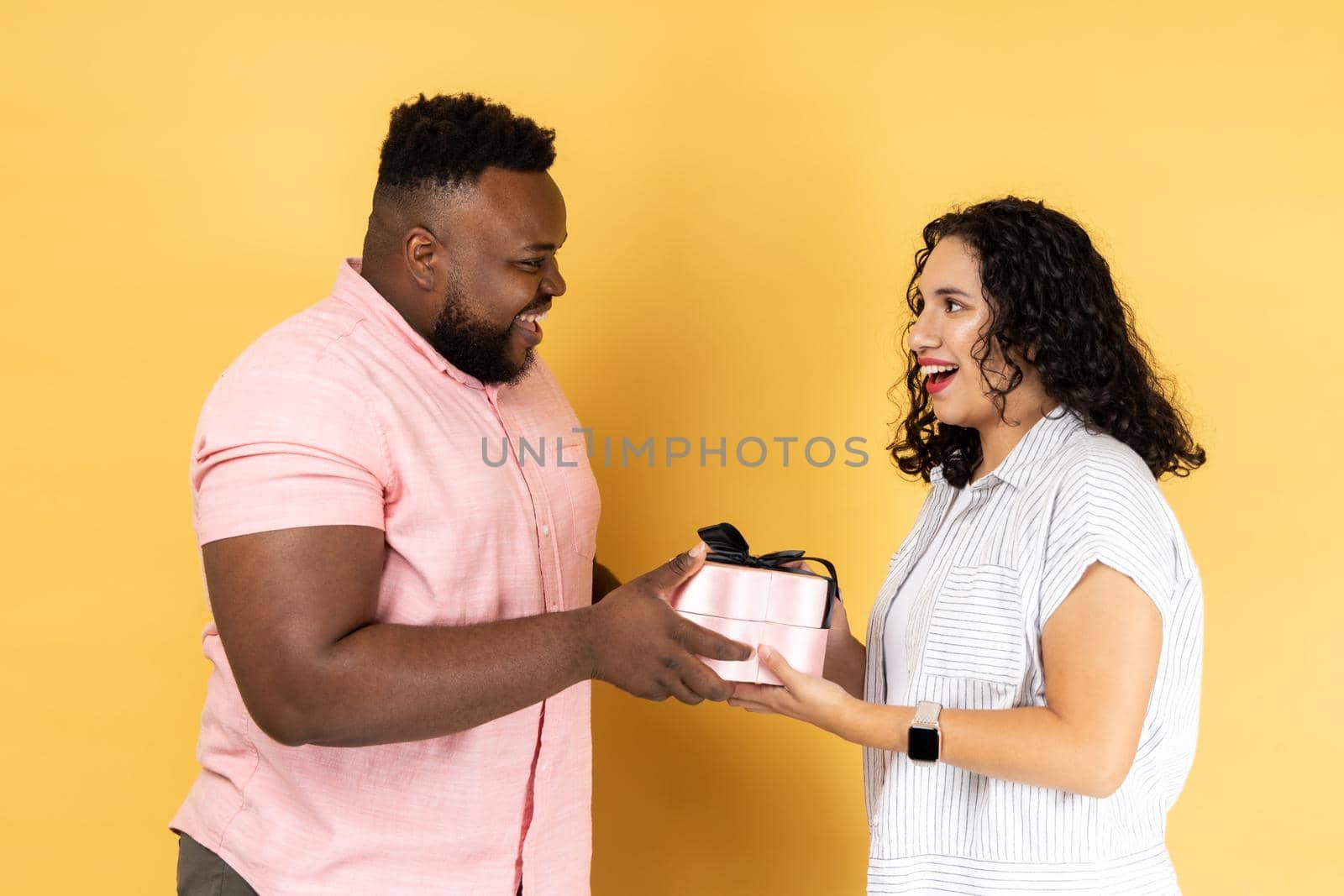 Portrait of couple in casual clothing standing together, celebrating anniversary, giving box with present to charming woman, looking at each other. Indoor studio shot isolated on yellow background.