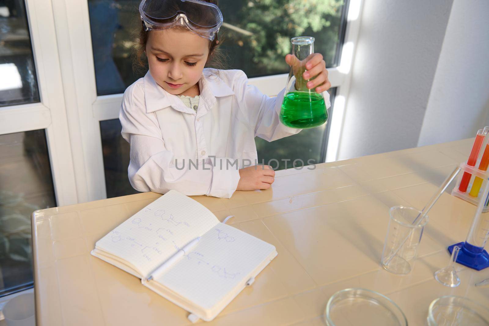 Charming little girl in lab wear, shaking a flask with green solution while reading the chemical formula in chemistry textbook in the school scientific laboratory. Back to school in new academic year