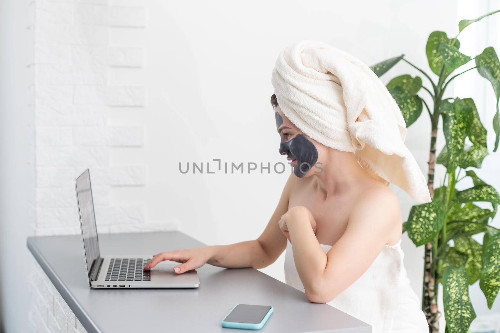 Young woman with towel on head and black facial clay mask sitting at home and using laptop computer . Staying at home, quarantine, distance learning, online education, technology, modern lifestyle.