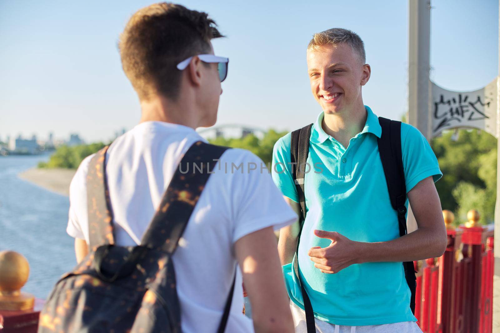 Outdoor portrait of two friends boys teenagers 15, 16 years old, talking laughing. Guys standing on bridge over river on sunny summer day. Youth, friendship, communication