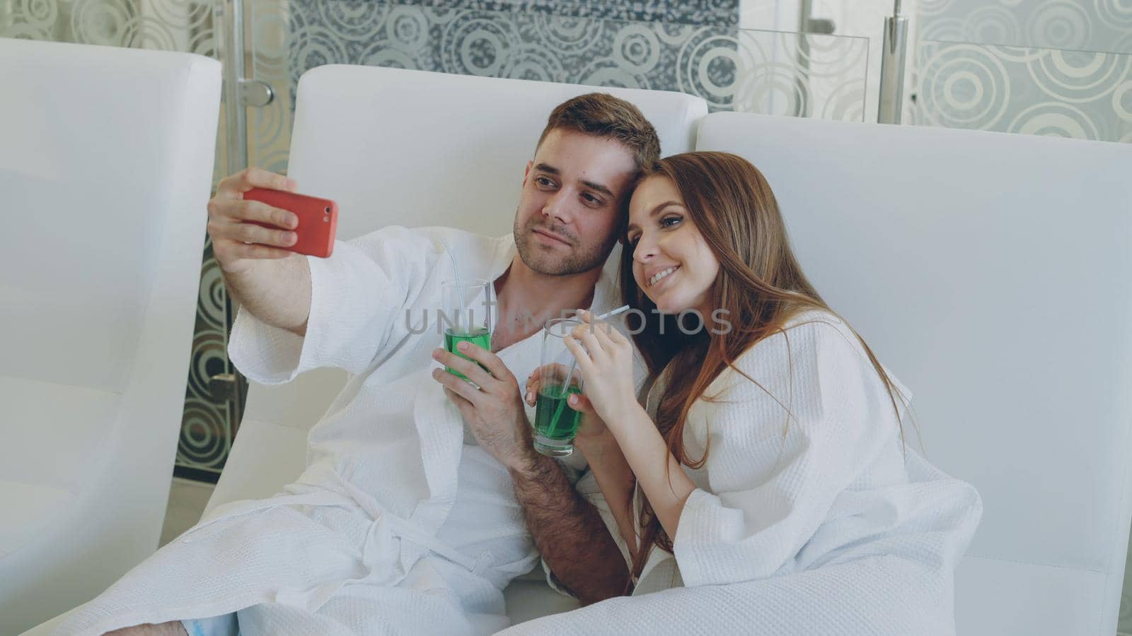 Attractive loving couple is taking selfie with cocktail glasses using smartphone while relaxing in spa salon. They are smiling and posing looking at camera. by silverkblack