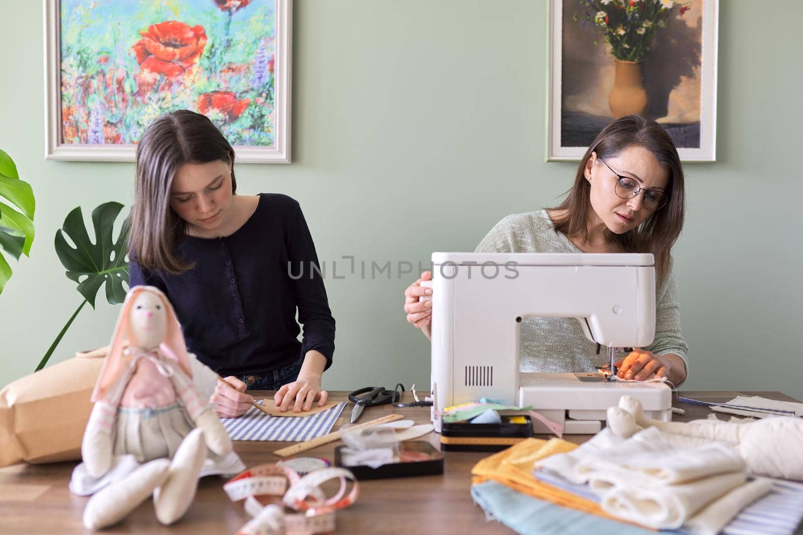 Family creative hand-made hobby and leisure, mother and teenager daughter together sew bunny toy doll. Woman teaches girl sewing skills, talking