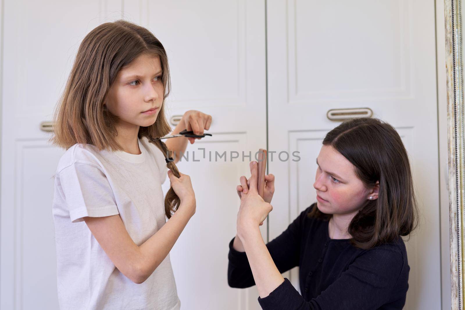 Girl child cuts her own hair with scissors, older sister teenager makes videos on smartphone, for social networks, blog, channel