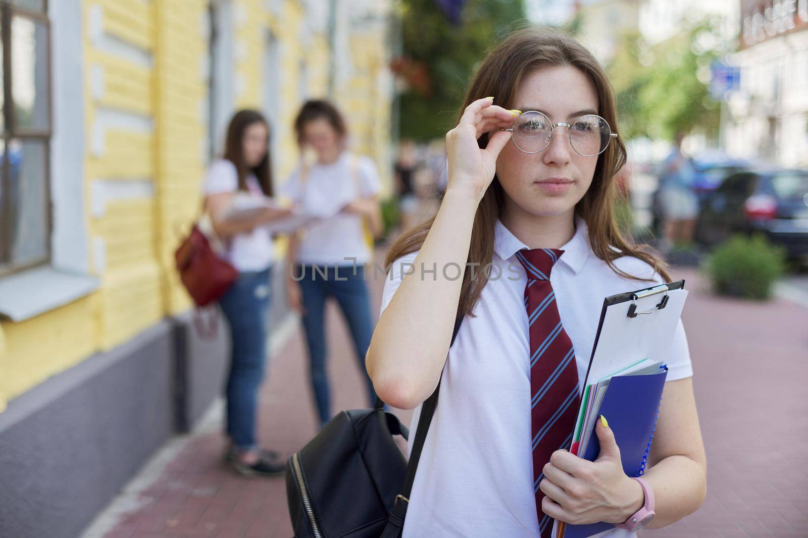 Portrait girl student teenager in glasses tie white T-shirt with backpack. Background yellow brick building, group of students