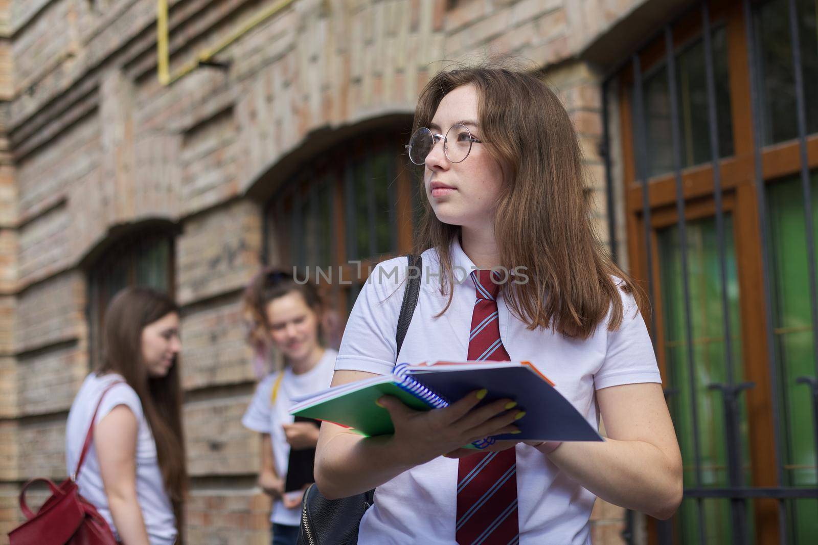 Girl teenager college student posing outdoor in white T-shirt with tie in glasses. Background brick building, group of girls students. Beginning of classes, back to college, copy space