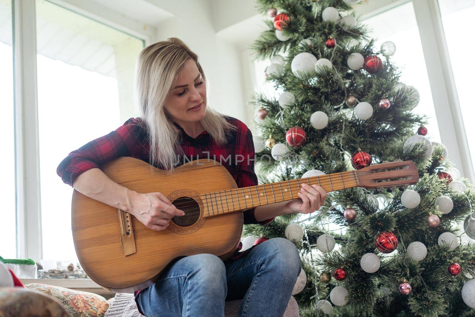 beautiful dark sits near the Christmas tree decorated with balls and light and playing the guitar and singing. Young beautiful smiling woman play guitar
