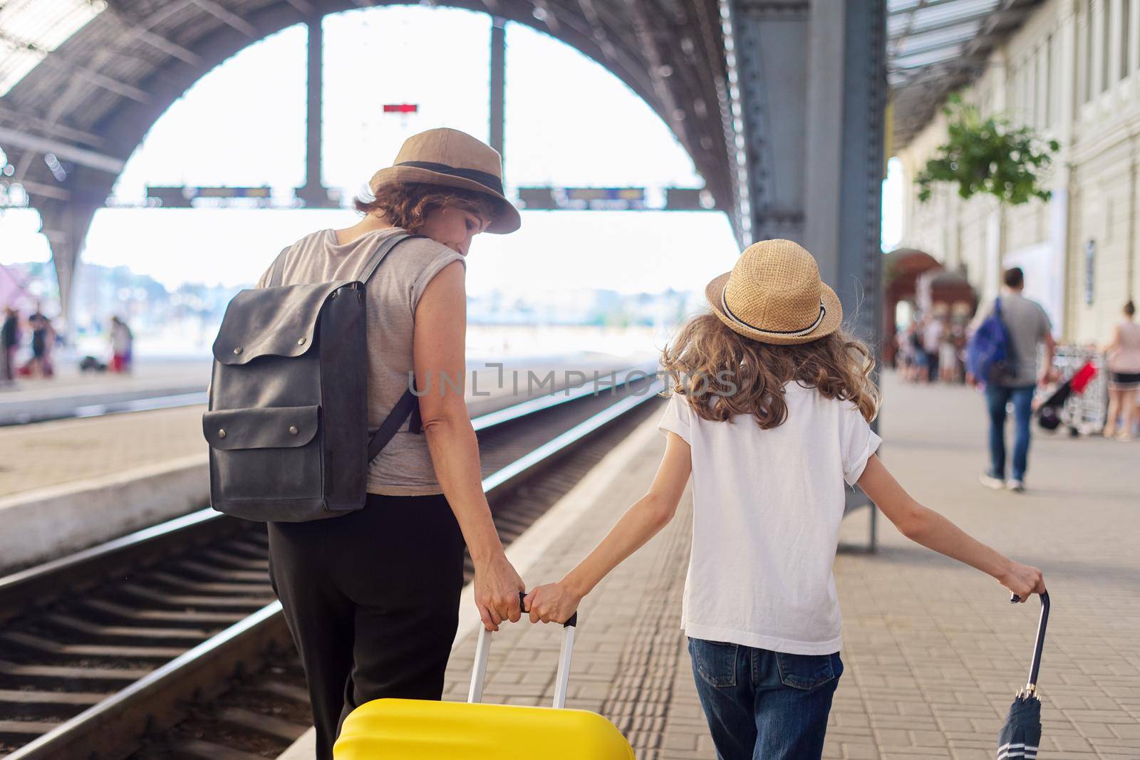 Happy mother and daughter child walking together at railway station with luggage suitcase, back view. Travel, tourism, transportation, family concept