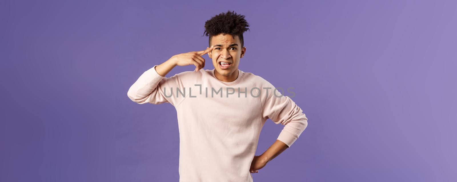 Portrait of angry, annoyed young man scolding someone from being stupid and crazy, rolling index finger over temple staring outraged and irritated camera, standing bothered purple background.