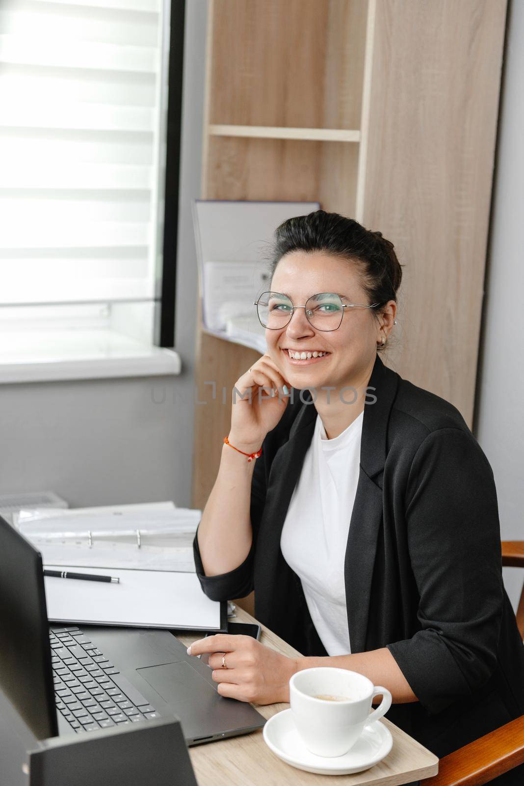 A young woman wearing glasses in a real office smiles and looks at the camera. Vertical photo. Hybrid work. Small office. by gulyaevstudio
