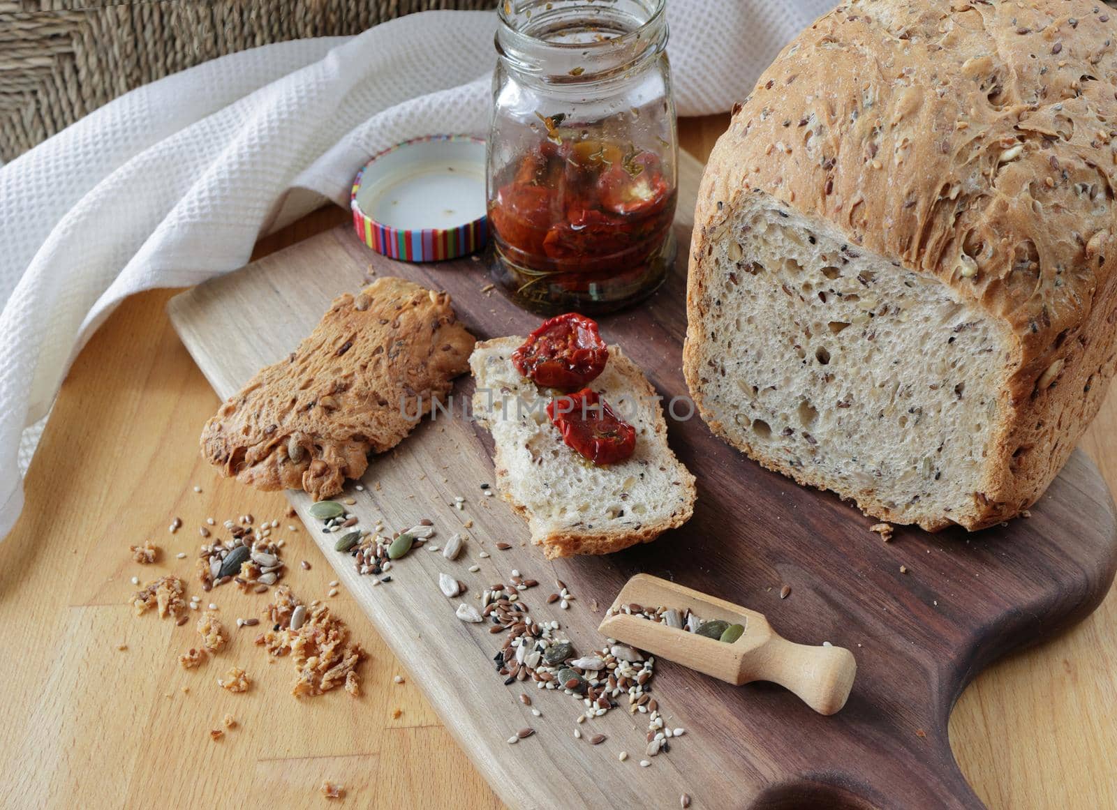 loaf of homemade whole grain bread and a cut slice of bread on a wooden cutting board. A mixture of seeds and whole grains. A jar of sun-dried tomatoes on a crust of fresh bread. Healthy food concept by Proxima13