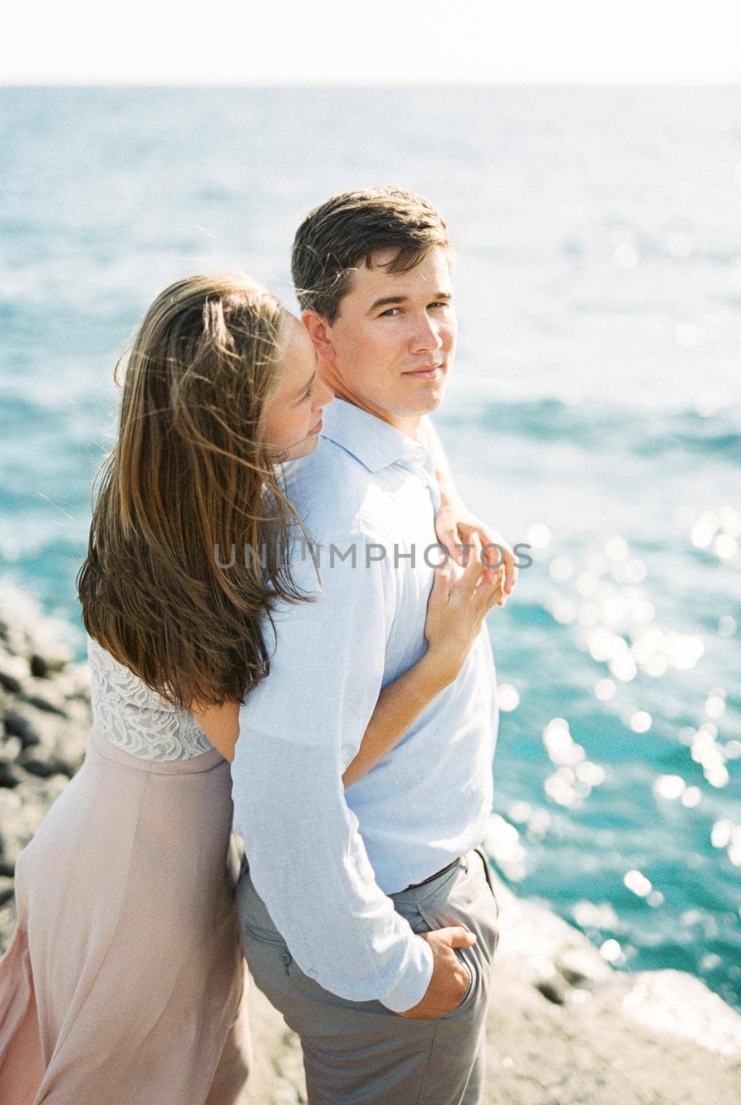 Young woman hugging a man from behind standing on a rock by the sea. High quality photo