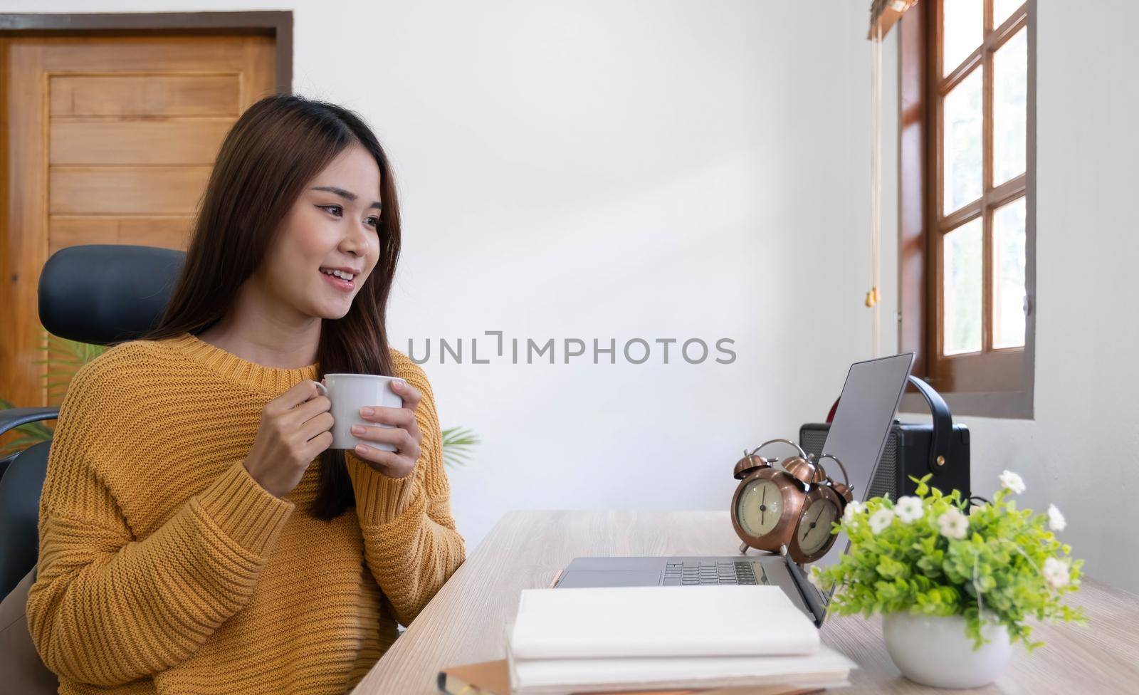 Beautiful young smiling asian woman working on laptop and drinking coffee in living room at home. Asia business woman working document finance and calculator in her home office. Enjoying time at home. by wichayada