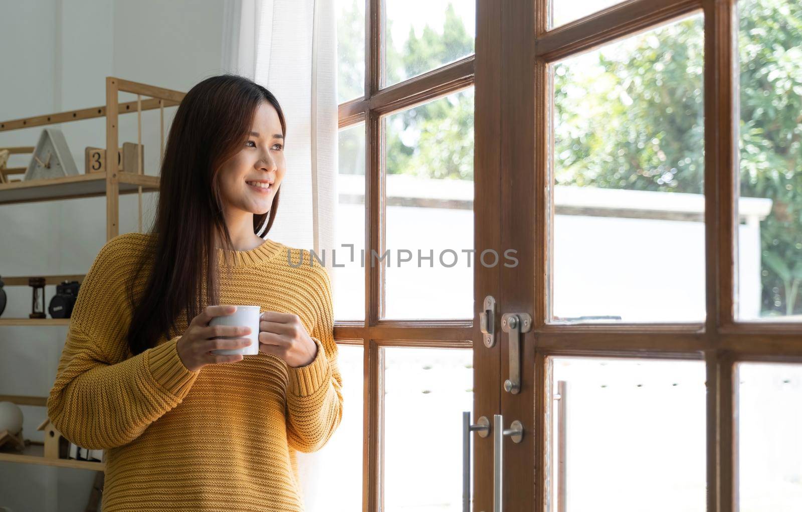 Happy young woman at home standing near window relaxing in her living room and drinking a cup of coffee..