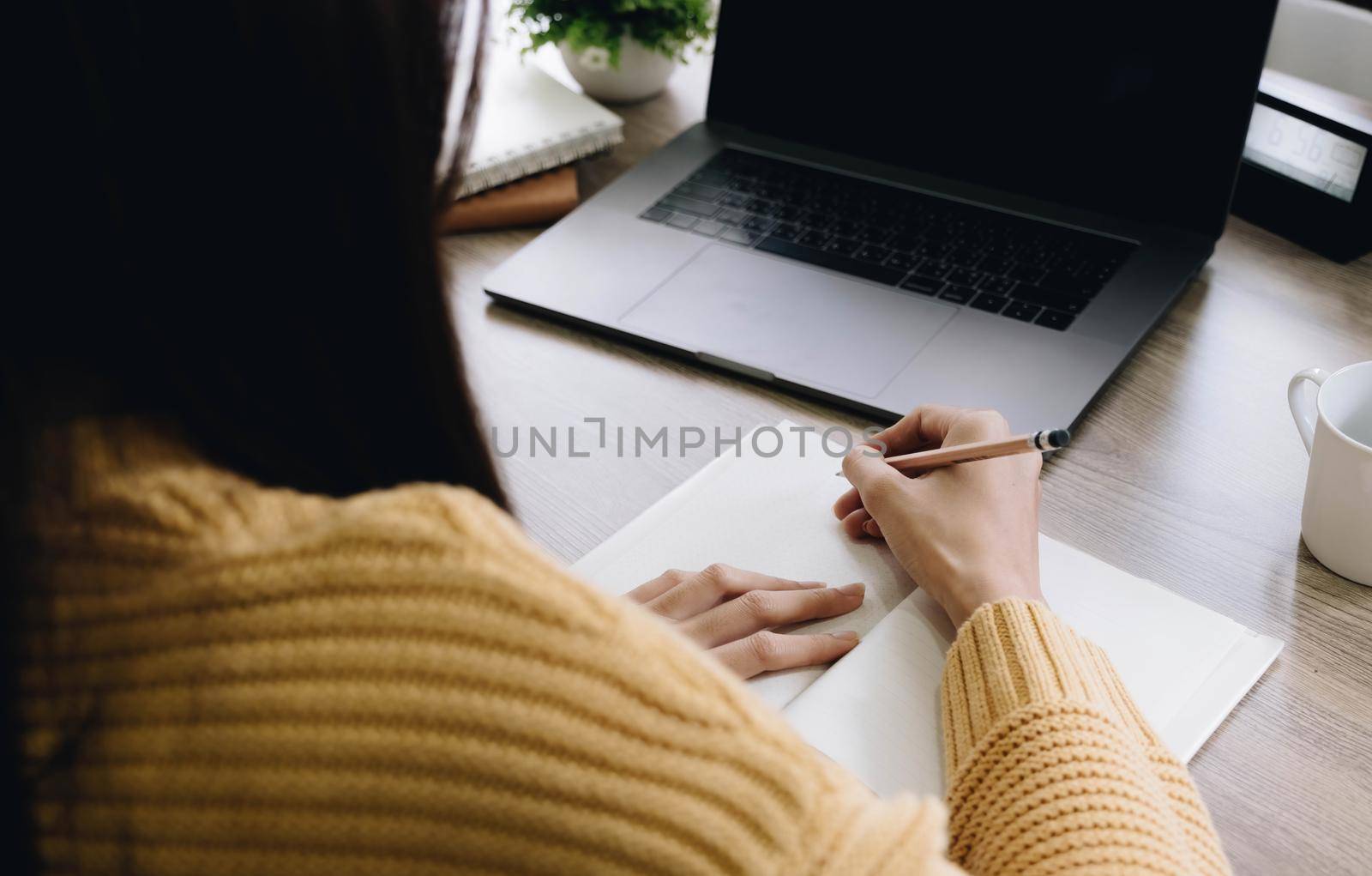 Beautiful woman writing taking notes while sitting in front her computer laptop at the wooden working table over living room bookshelf as background..