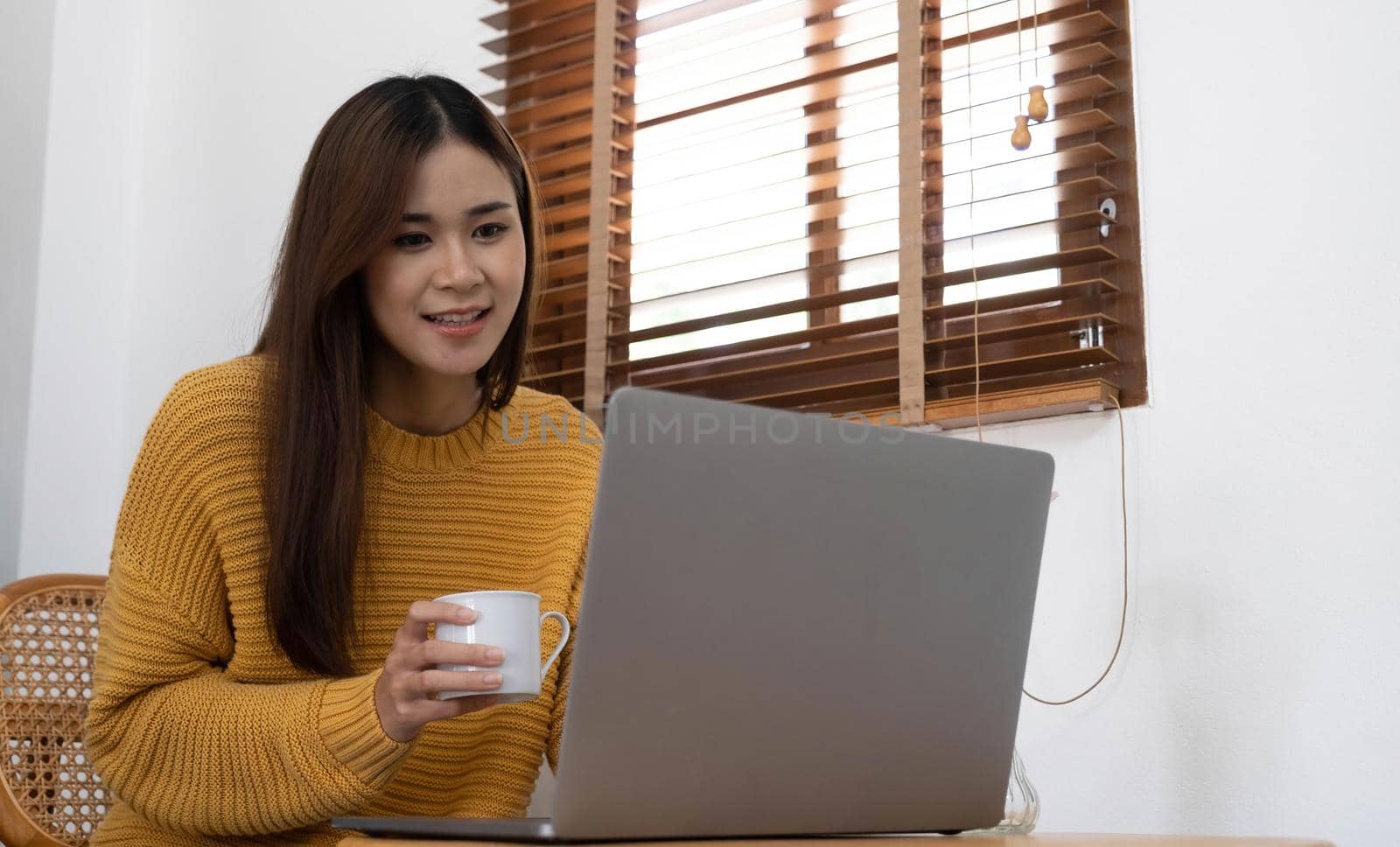 Young woman relaxing and drinking cup of hot coffee or tea using laptop computer on chair.woman checking social apps and working.Communication and technology concept by wichayada