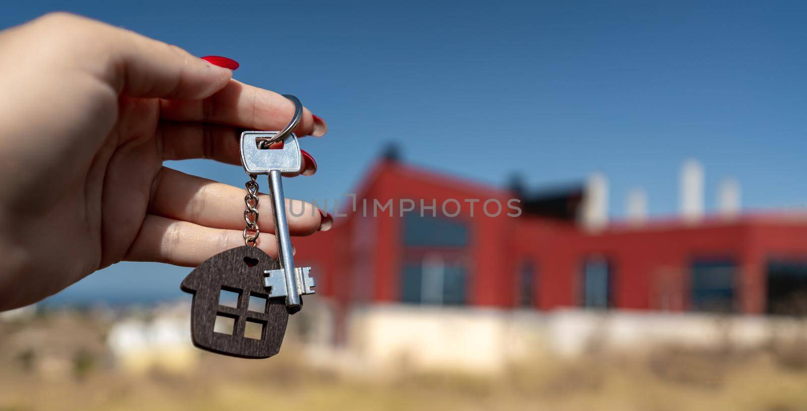 A woman's hand holds a house key against the backdrop of a house under construction. Real estate agent. Buying a house, apartment