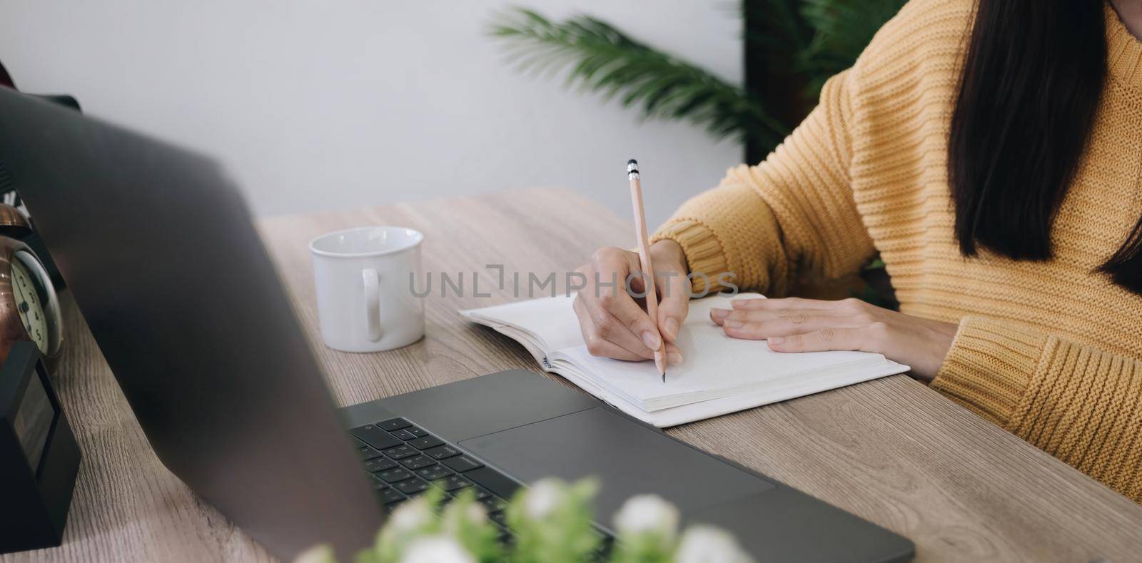 Beautiful woman writing taking notes while sitting in front her computer laptop at the wooden working table over living room bookshelf as background. by wichayada