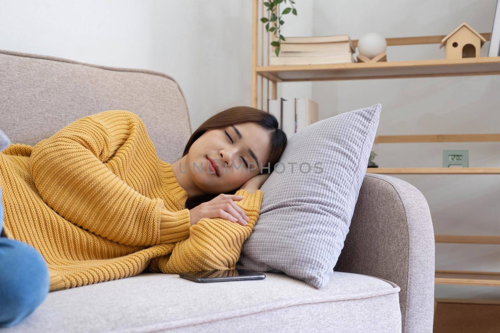 beautiful asian woman in sweater sleeping on a couch at home with mobile phone in home by wichayada