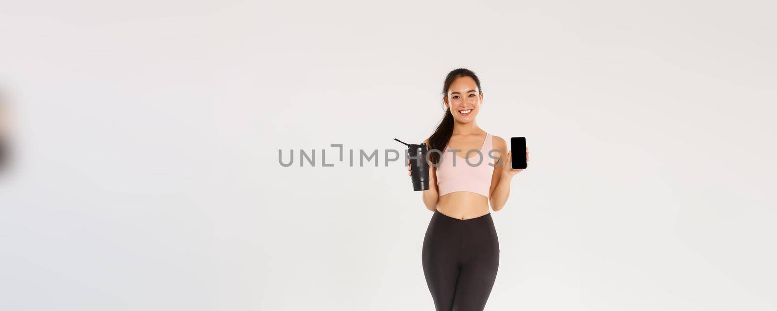 Full length of smiling good-looking asian fitness girl, sportswoman in activewear showing smartphone screen and water bottle, control drinking liquid with mobile application during training session.