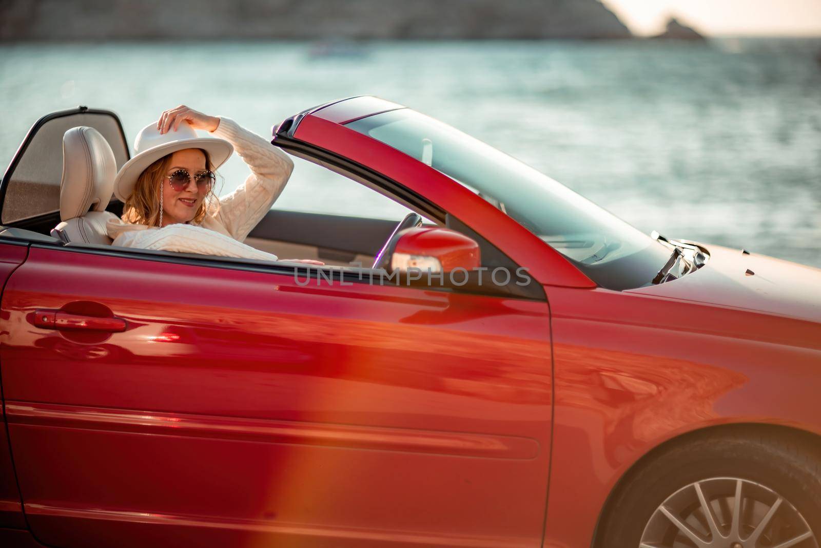 Outdoor summer portrait of stylish blonde woman driving red car convertible. Fashionable attractive woman with blond hair in a white hat in a red car. Sunny bright colors taken outdoors against the sea