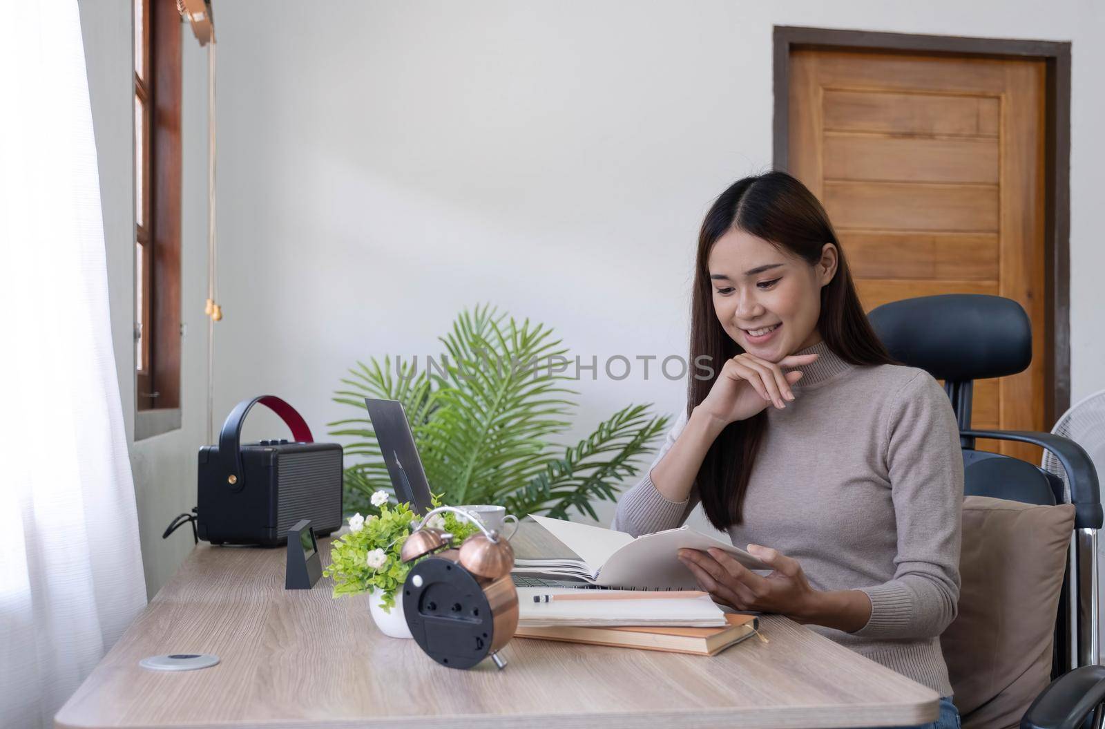 Attractive young woman sits in a chair and looks at a book in her hand. girl on the right There is a laptop on the desk in the office. by wichayada