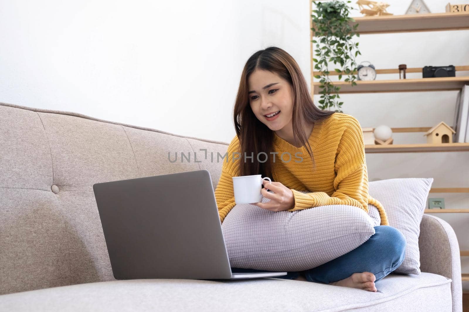 Young asian business woman work from home with laptop computer and drinking coffee on sofa in living room, freelance girl using notebook sitting on couch with comfort and relax, lifestyles concept..