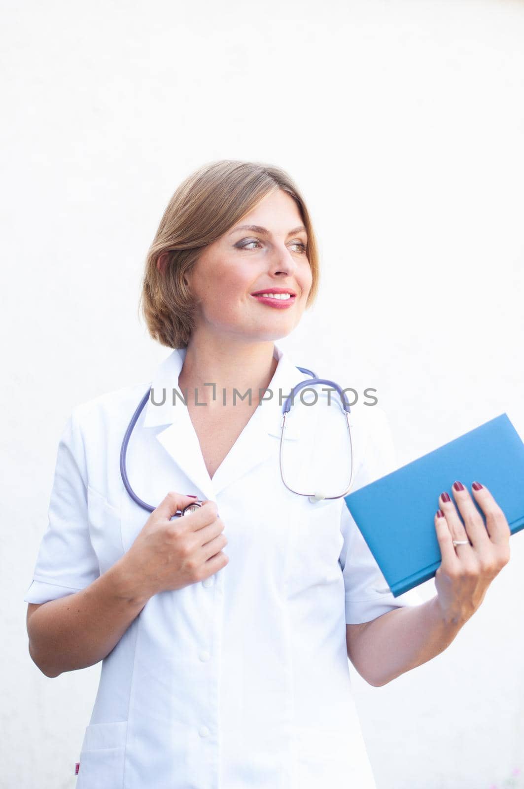 Young beautiful caucasian woman doctor with a phonendoscope on her neck and with a notepad smiling on a light background. High quality photo
