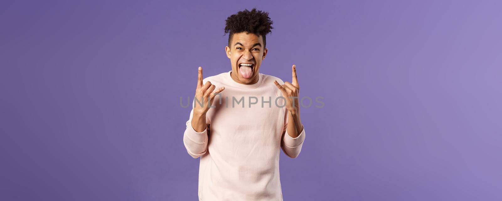 Portrait of enthusiastic, excited funny young man having fun, enjoying awesome rock concert, show heavy metal sign, stick tongue and rejoicing hear favorite band on stage, purple background.