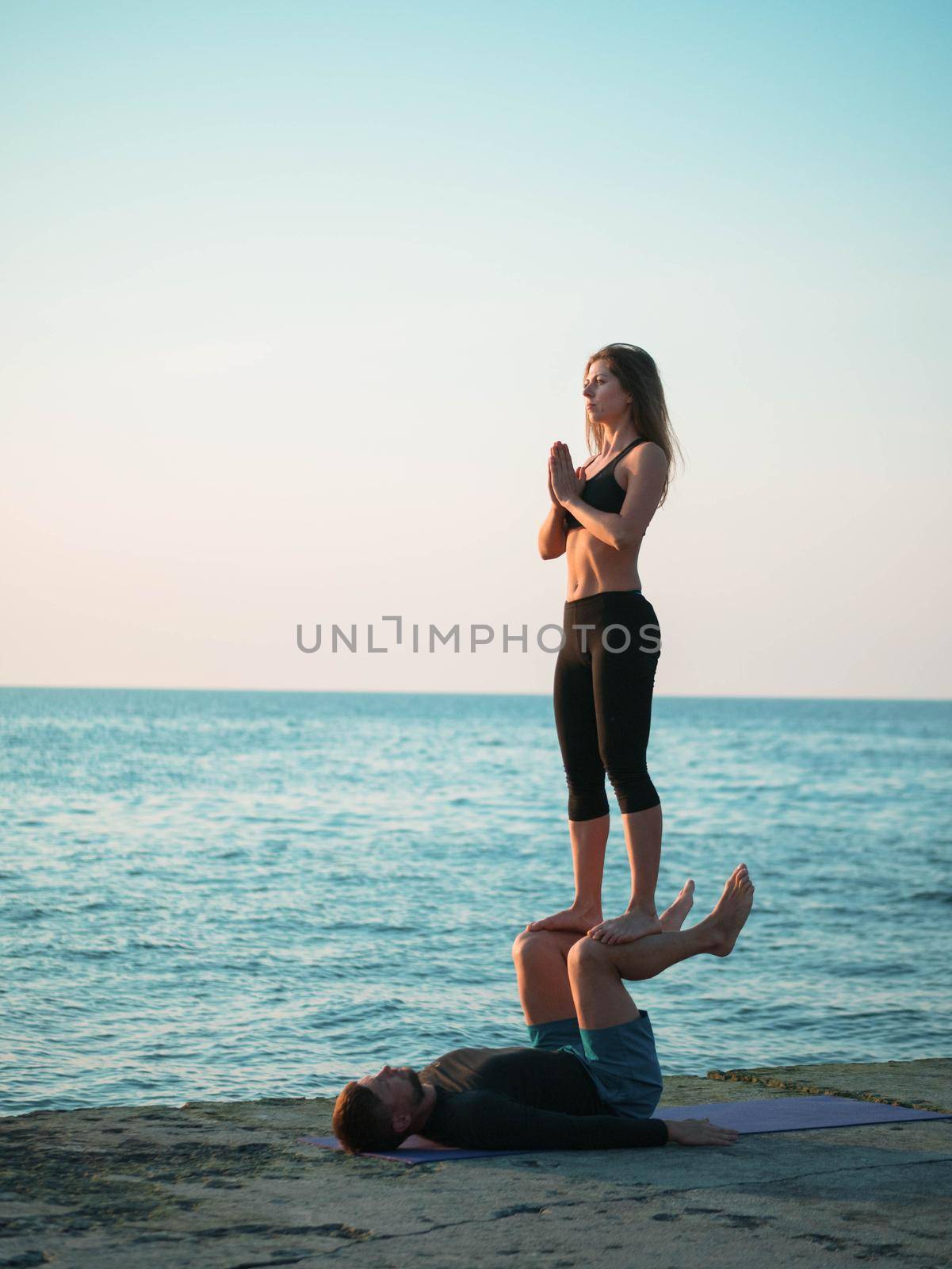 Young beautiful couple practicing acro yoga on the sea beach near water. Man and woman doing everyday practice outdoor on nature background. Healthy lifestyle concept.