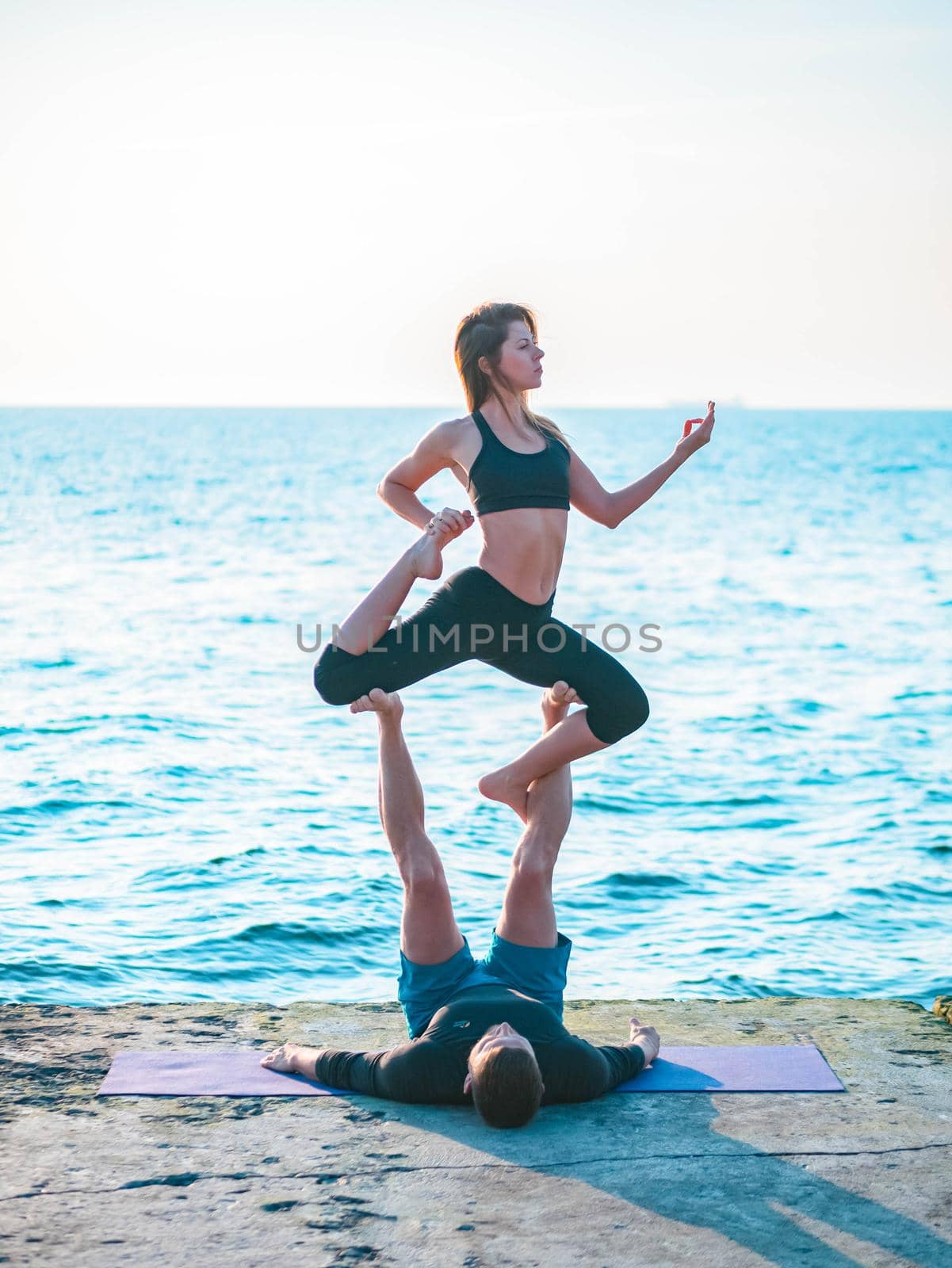 Young beautiful couple practicing acro yoga on the sea beach near water. Man and woman doing everyday practice outdoor on nature background. Healthy lifestyle concept. by kristina_kokhanova
