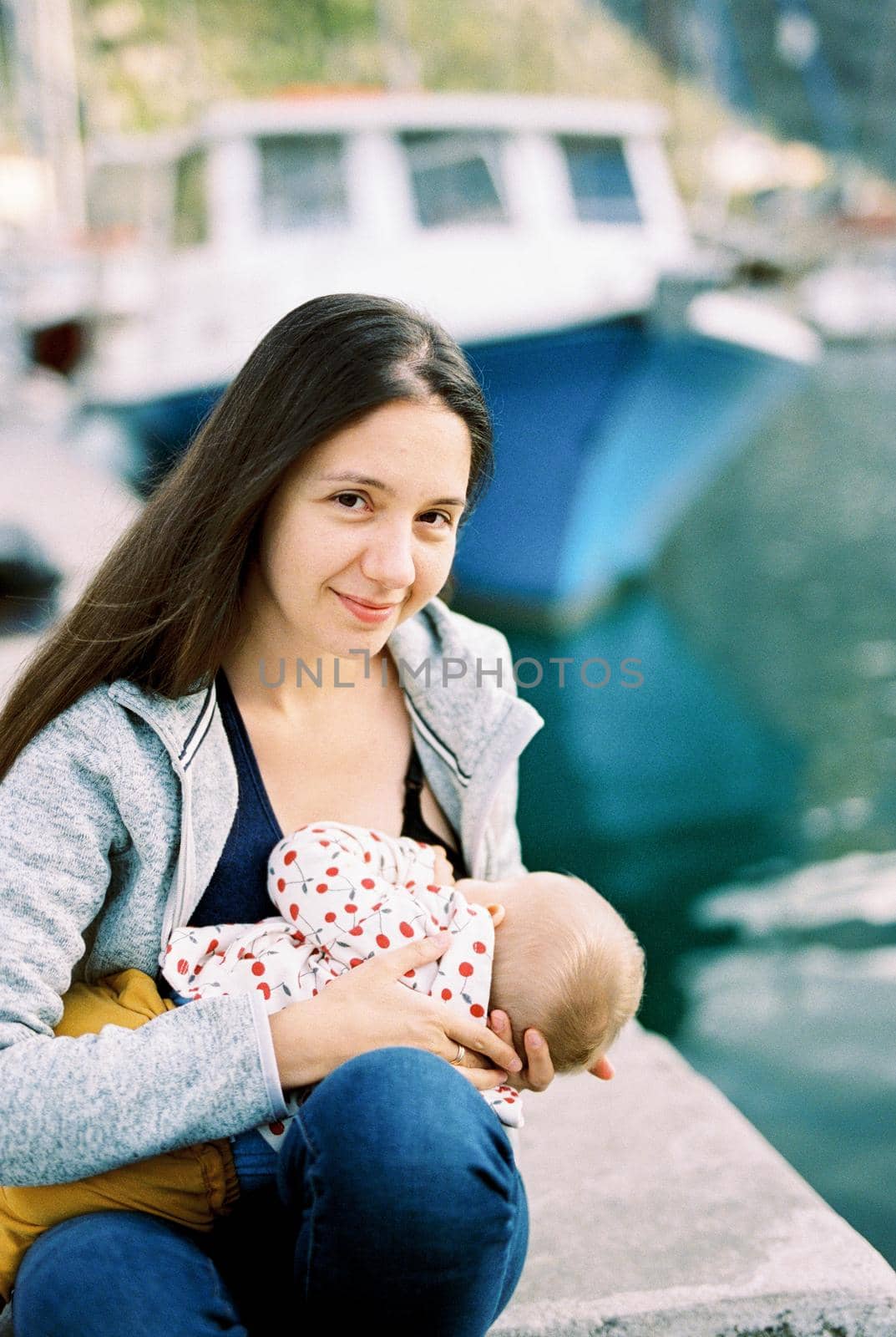 Mom breastfeeds her baby while sitting on the pier by Nadtochiy