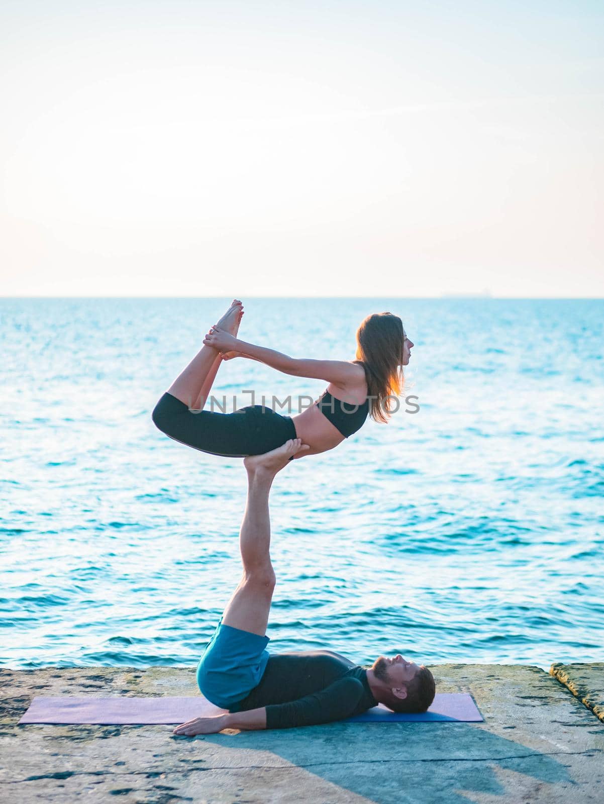 Young beautiful couple practicing acro yoga on the sea beach near water. Man and woman doing everyday practice outdoor on nature background. Healthy lifestyle concept. by kristina_kokhanova