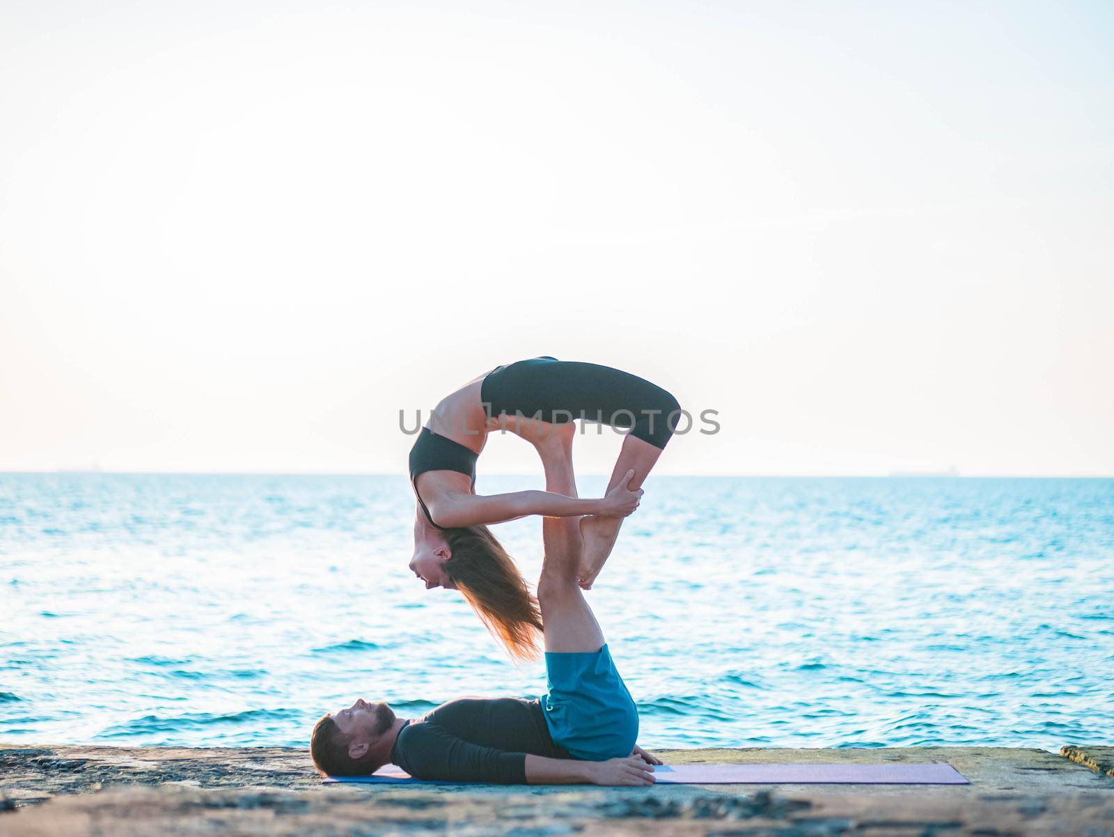 Acroyoga concept. Two sporty people practicing yoga in pair on nature sunrise background. Beautiful young couple doing stretching exercise on the sea beach. by kristina_kokhanova