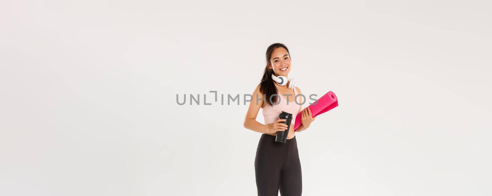 Full length of smiling slim and beautiful asian female athlete, girl carry fitness exercise rubber mat and water bottle, going for yoga classes or workout in gym with own equipment, white background.
