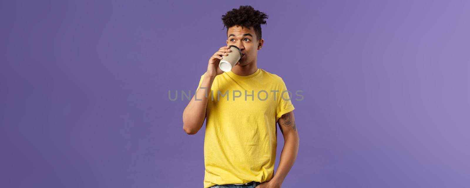 Portrait of carefree young male student getting energy in morning from caffeine, sipping coffee from take-away cup, look away casually standing, order drink in cafe, purple background by Benzoix