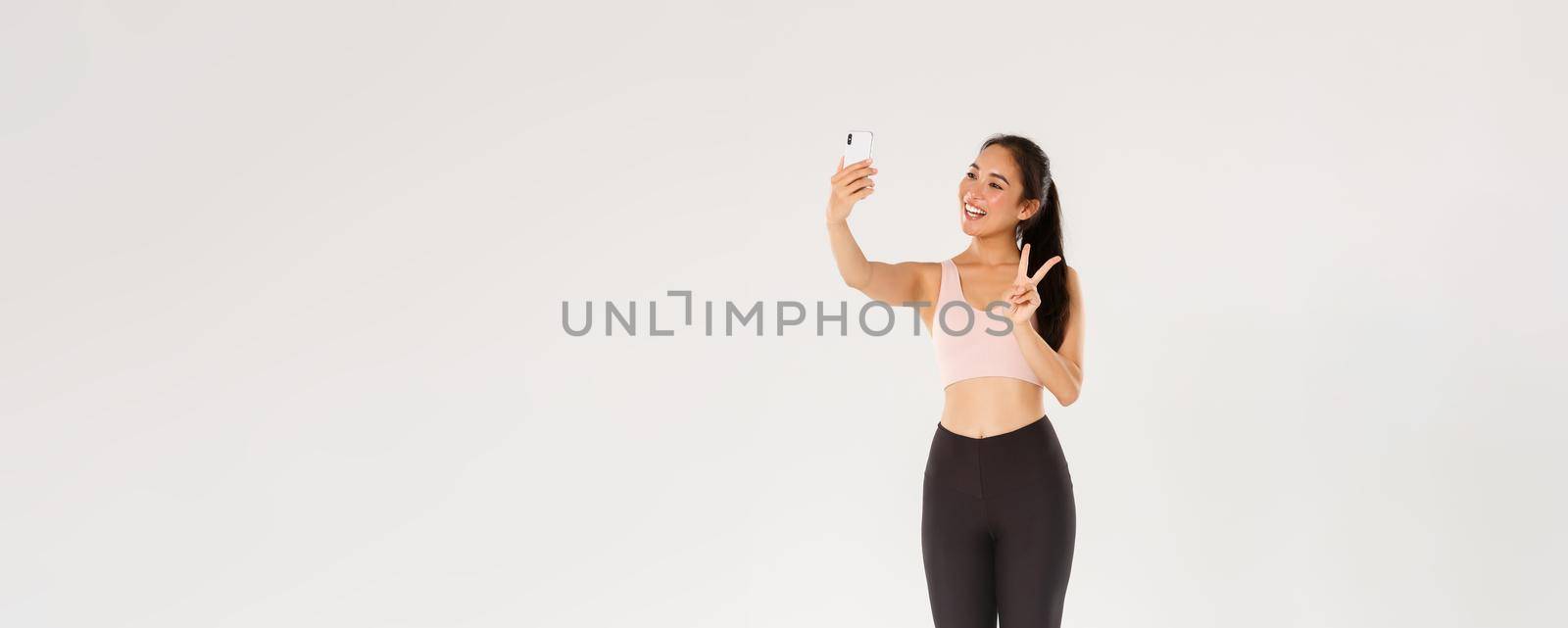 Full length of cute asian fitness girl, sportswoman in sportswear showing peace gesture and taking selfie on mobile phone, woman workout in gym and taking pics of herself during training session.
